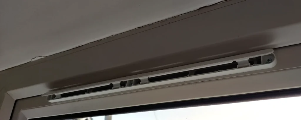 An open trickle vent: a slim white plastic vent with slots, fitted along the top of a uPVC window frame.